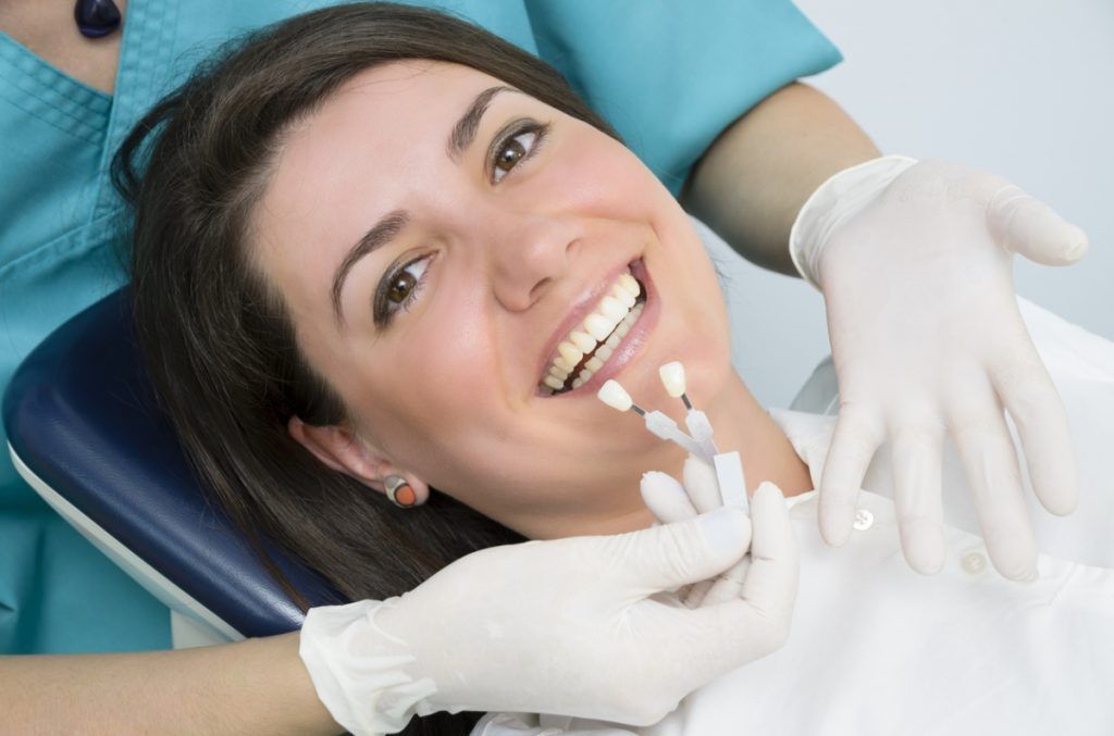 A woman is sitting in a dental chair smiling while a dentist examines her teeth.