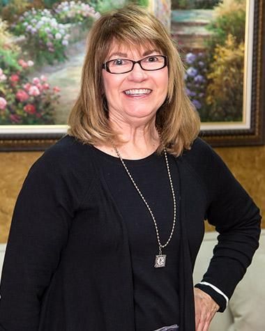 A woman wearing glasses and a necklace is standing in front of a painting.