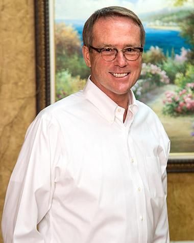 A man wearing glasses and a white shirt is standing in front of a painting.