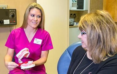 A woman is sitting in a dental chair talking to a nurse who is holding a model of teeth.