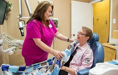 A woman in a pink shirt is talking to an older woman in a dental chair.