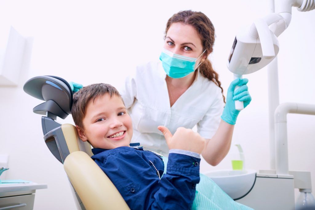 A young boy is sitting in a dental chair giving a thumbs up.