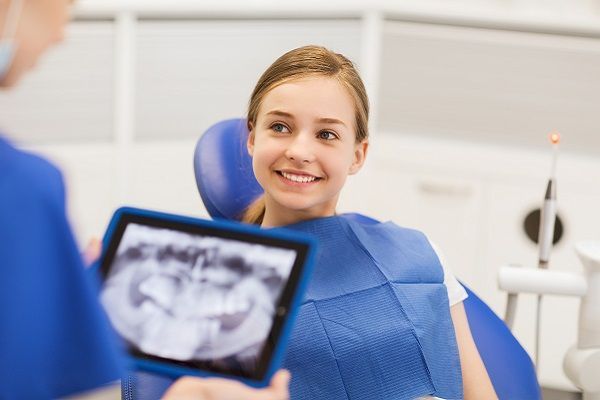 A young girl is sitting in a dental chair looking at an x-ray of her teeth.