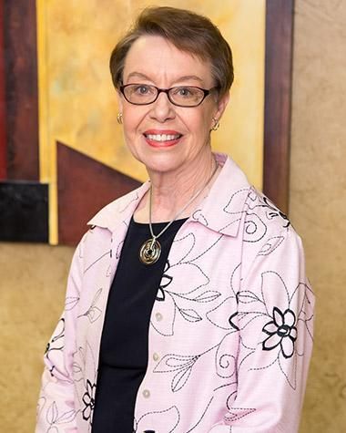 A woman wearing glasses and a pink jacket is smiling in front of a painting.