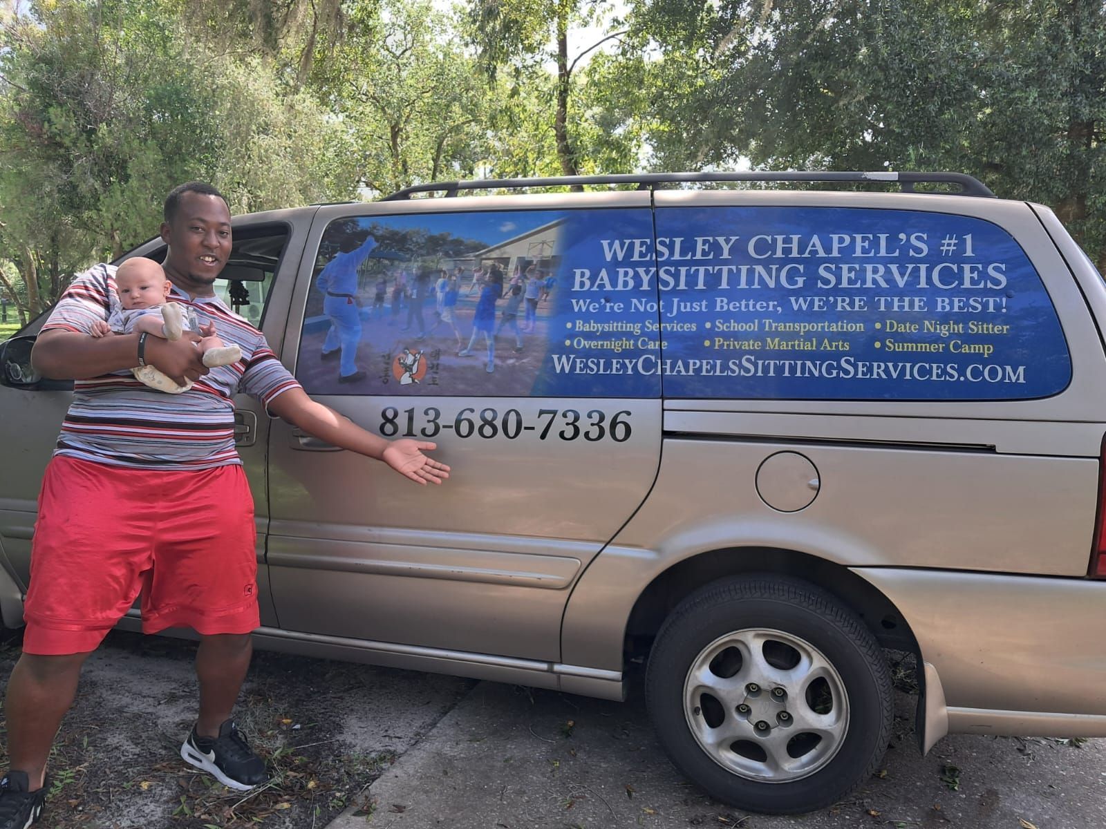 A man is holding a baby in front of a van that says wesley chapel 's # 1 babysitting services