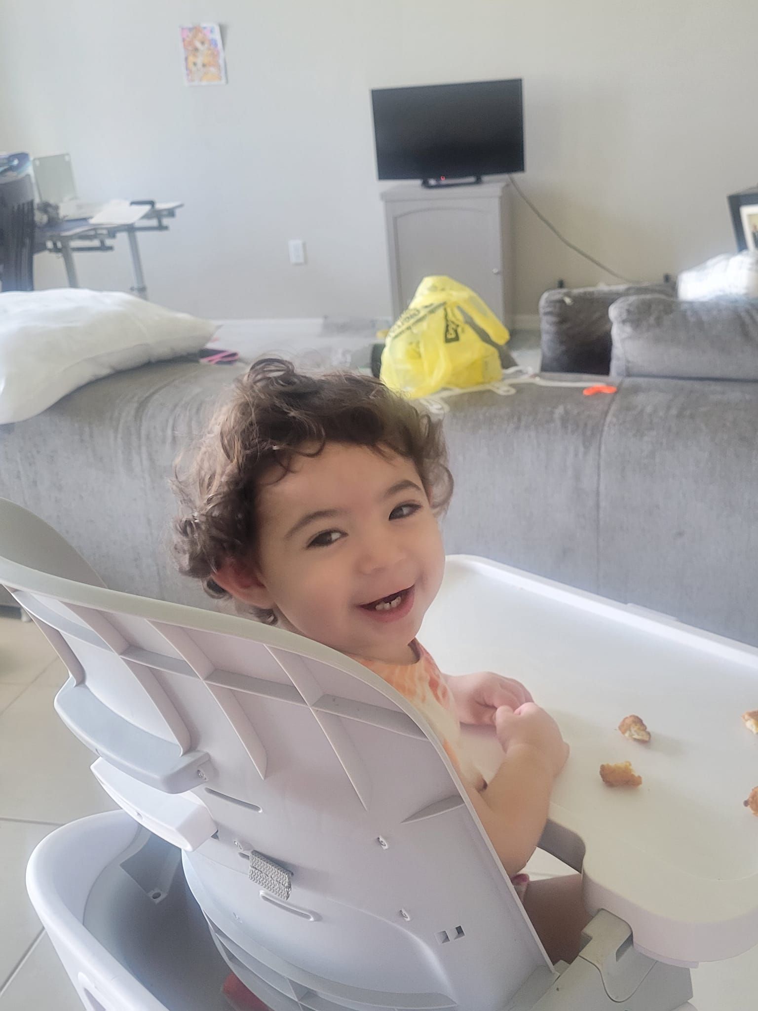 A baby is sitting in a high chair in a living room and smiling.