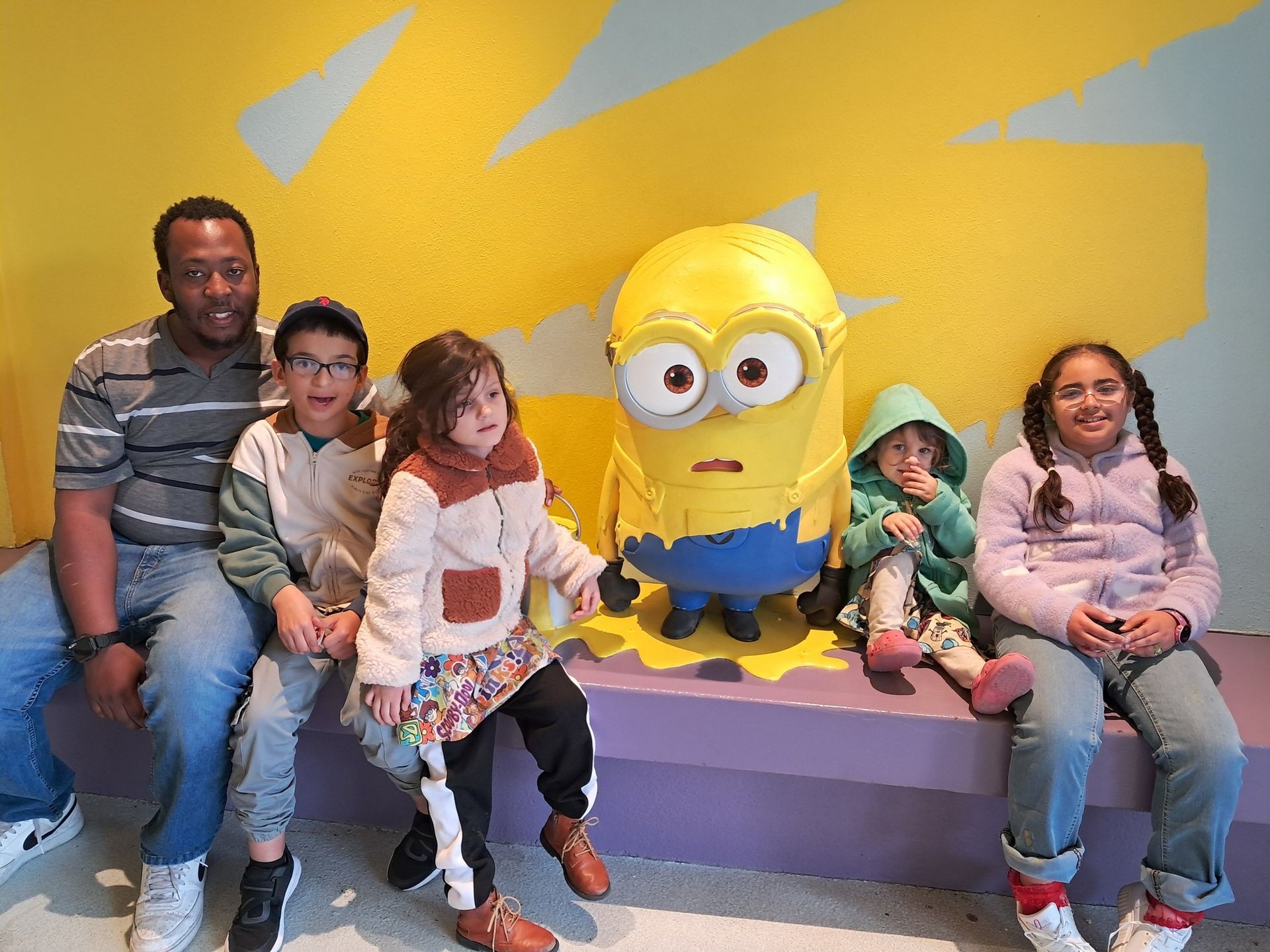 A group of children are sitting next to a statue of a minion.