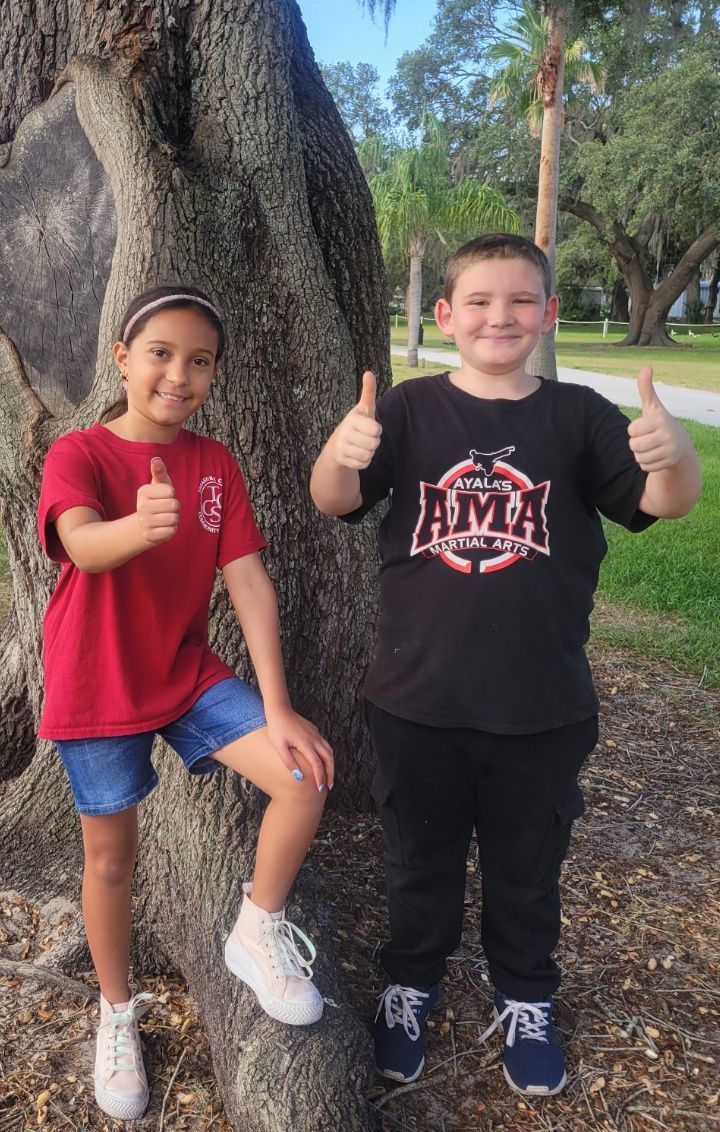 A boy and a girl are standing next to a tree giving a thumbs up.