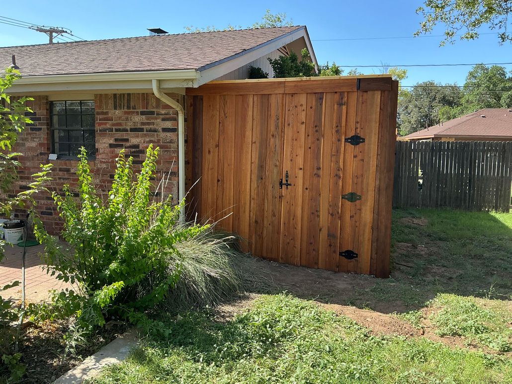 A tall, vertical wood privacy fence stands attached to the side of a brick house next to a small green bush.