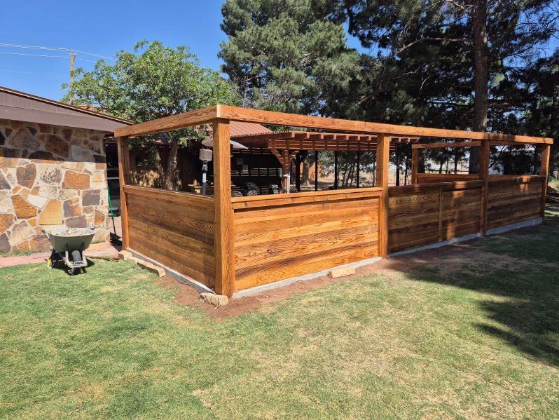 A wooden fence and pergola structure stands in a backyard, adjacent to a stone-walled building and grass lawn.