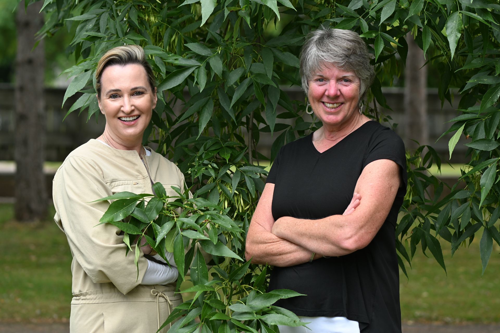 Two women smiling outdoors; one in tan jumpsuit, the other in black shirt. Lush green foliage surrounds them.