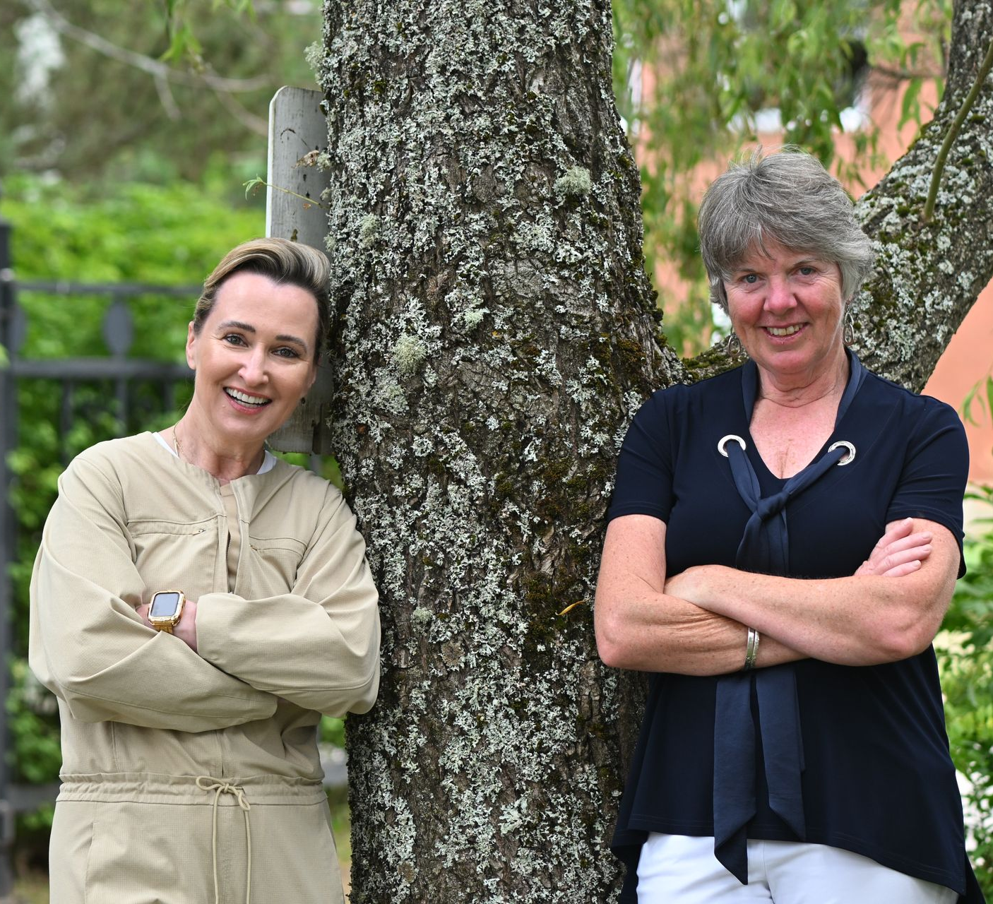 Two women stand next to a tree, arms crossed. One wears a tan jumpsuit, the other, a black top and white pants. Outdoors.