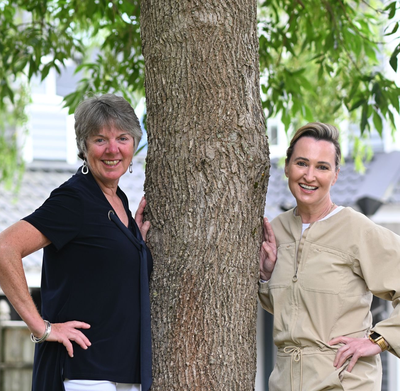 Two women standing next to tree, smiling. One wears black shirt, the other a beige jumpsuit, in a yard.