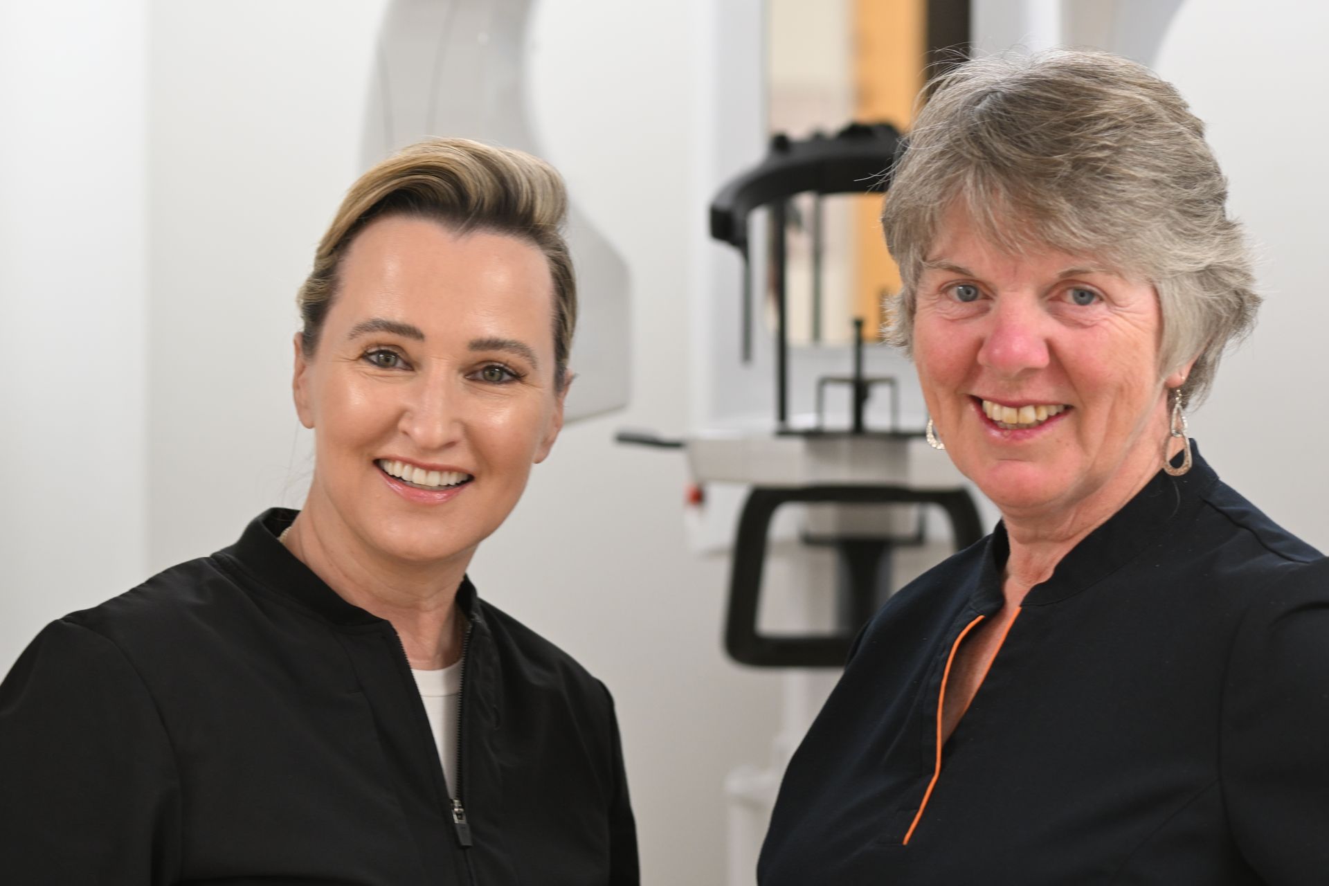 Two women smiling, dressed in black tops, standing in a dental office next to X-ray equipment.
