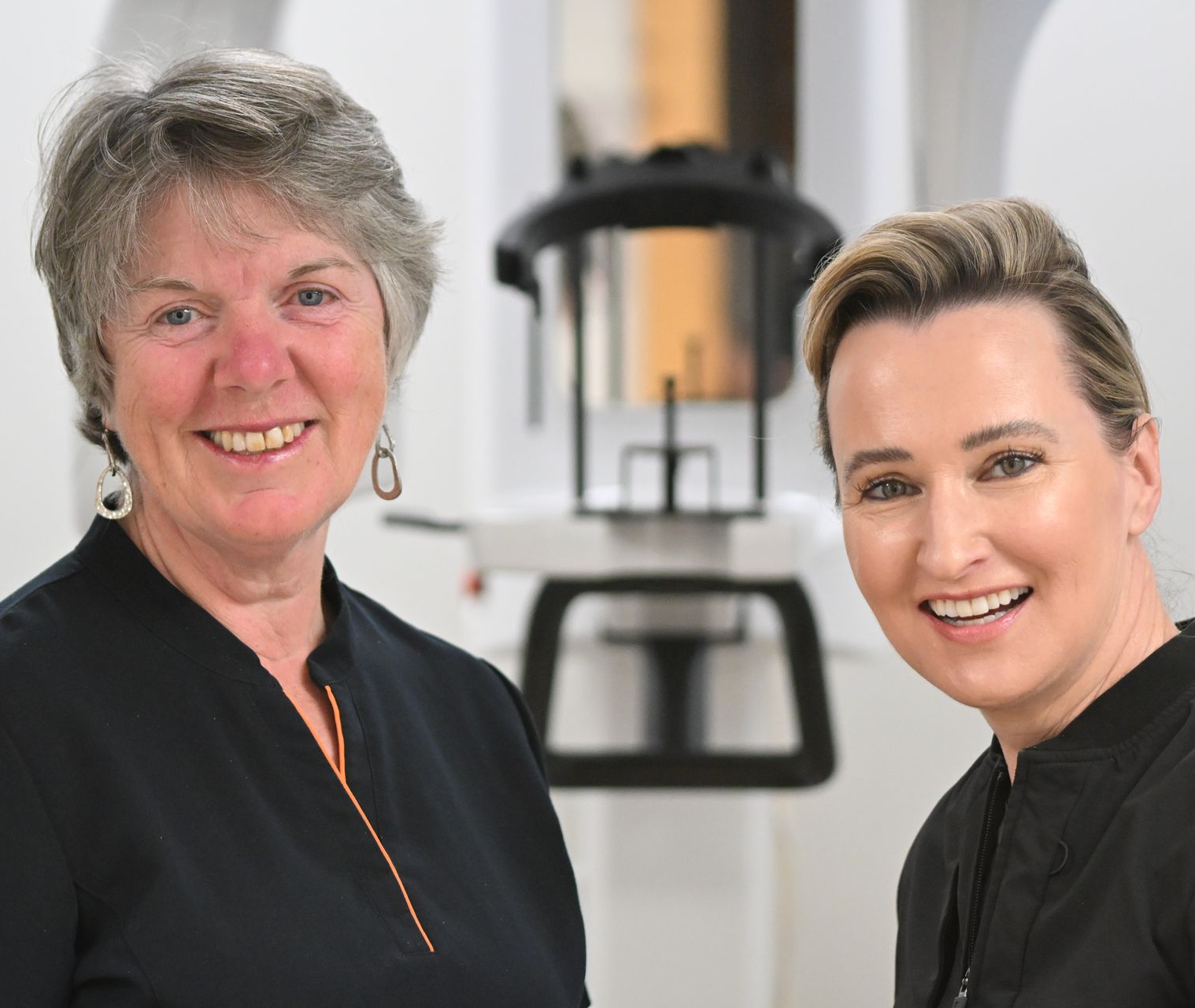 Two smiling women in black tops pose in front of dental equipment.