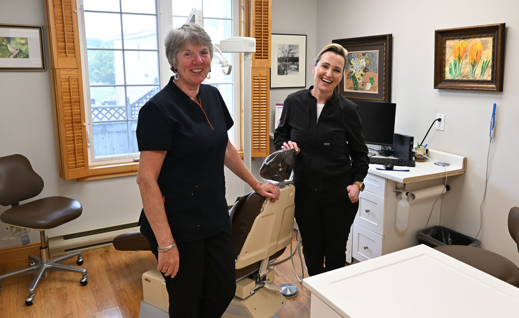 Woman with gray hair smiles, wearing a black shirt, standing in a dental office.