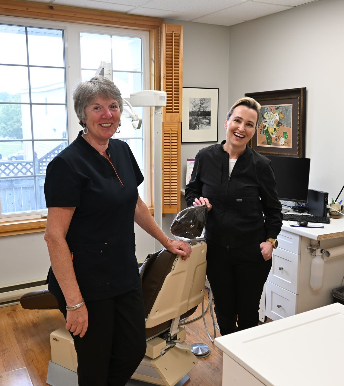 Two women in black scrubs in a dental office. One by a dental chair, the other by a desk. Both smiling.