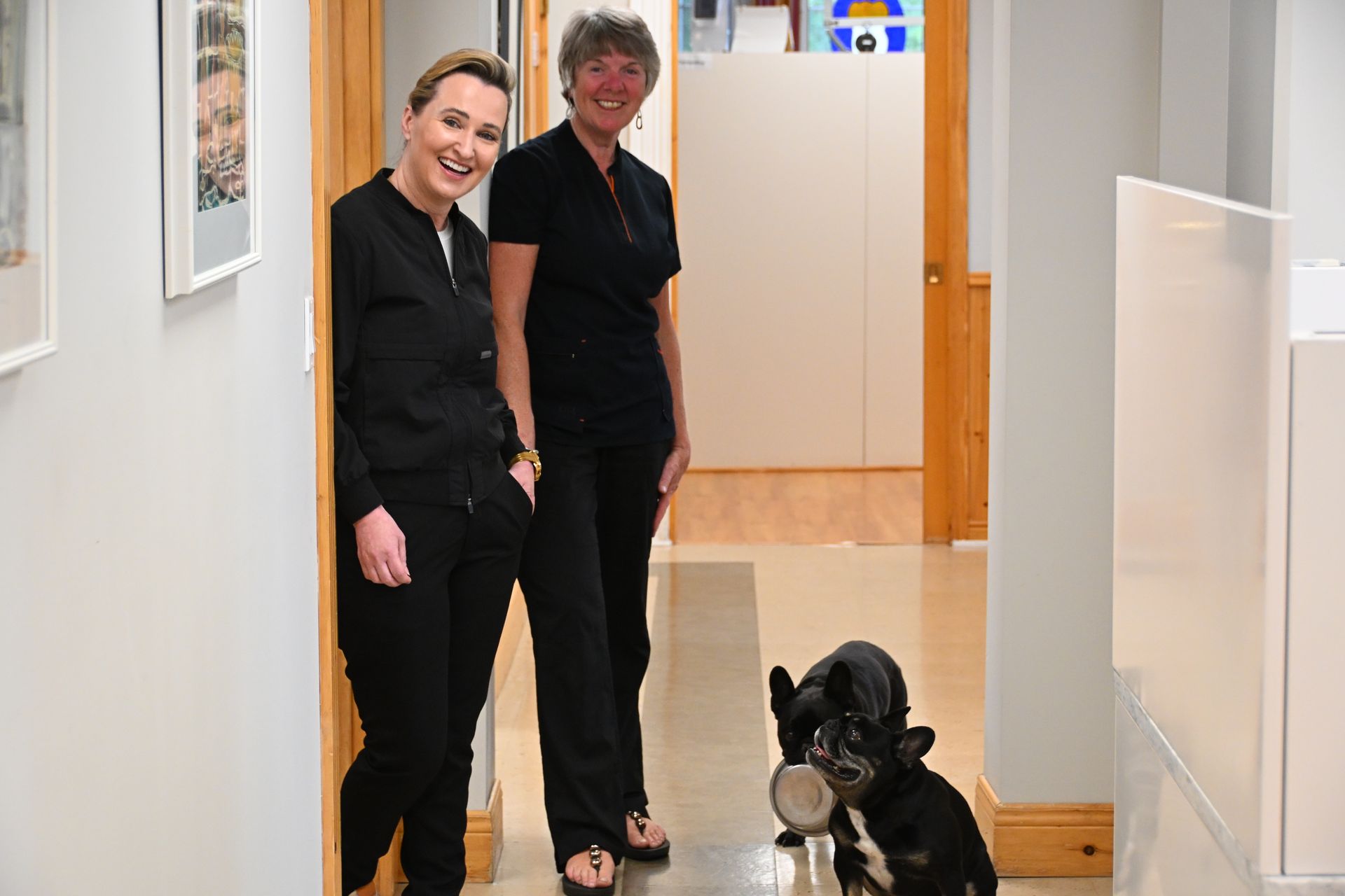 Two women in black uniforms smile in a hallway; a dog holds a bowl.