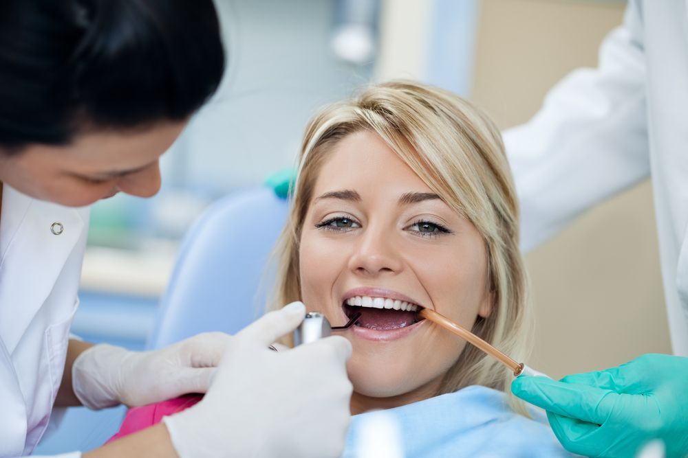 Woman in dental chair, mouth open; dentists examining her teeth with tools.