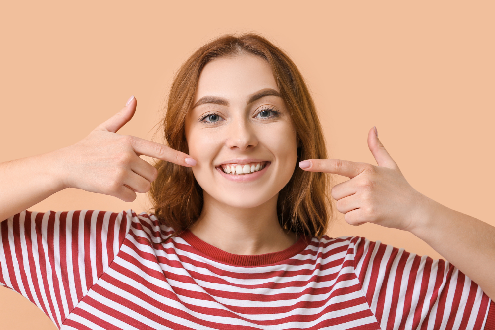 Woman with red hair smiling, pointing to her bright white teeth.