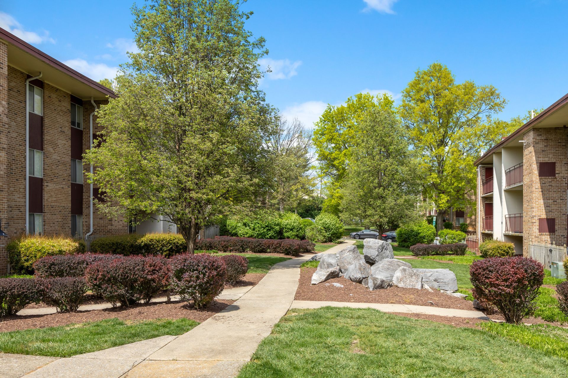 A brick apartment building with a walkway leading to it surrounded by trees and bushes.