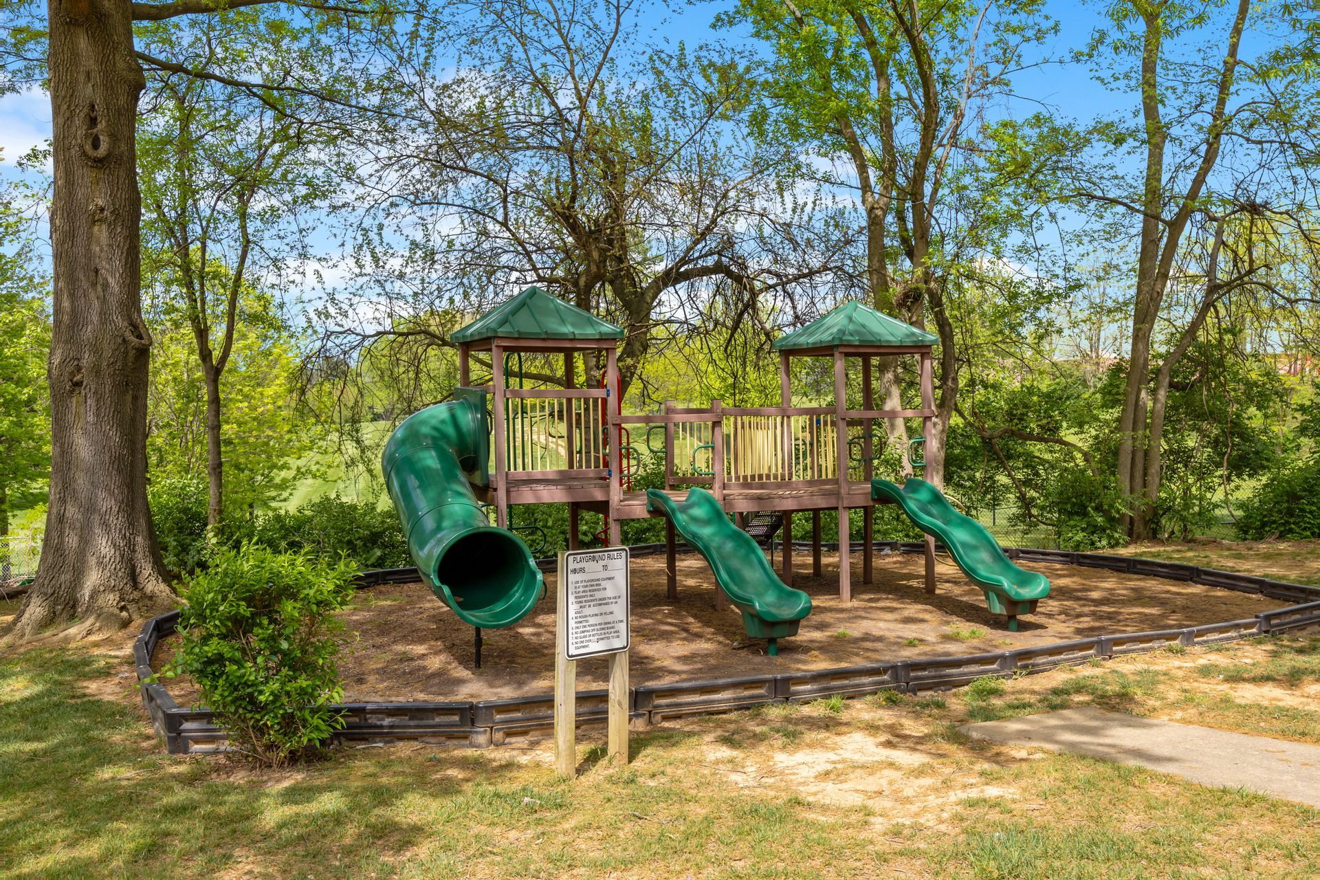A wooden playground with a green slide in the middle of a park surrounded by trees.