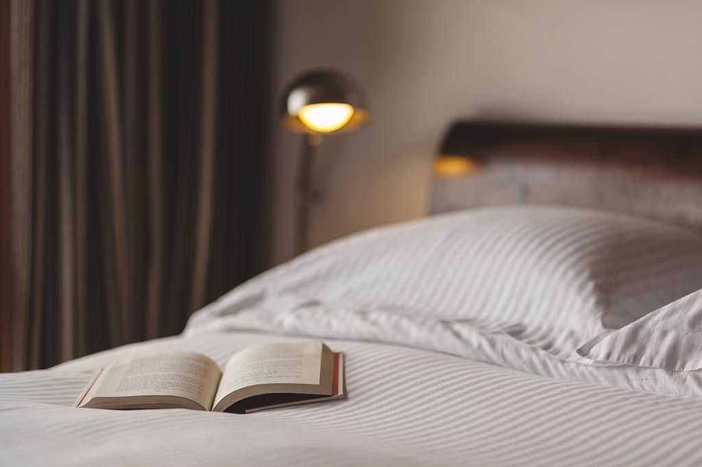 Open book on a bed with white and gray striped linens; lamp and curtains in the background.