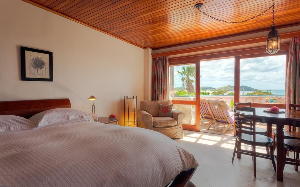 Bedroom with wood panel ceiling, large bed, art, dining table, and balcony view at night.