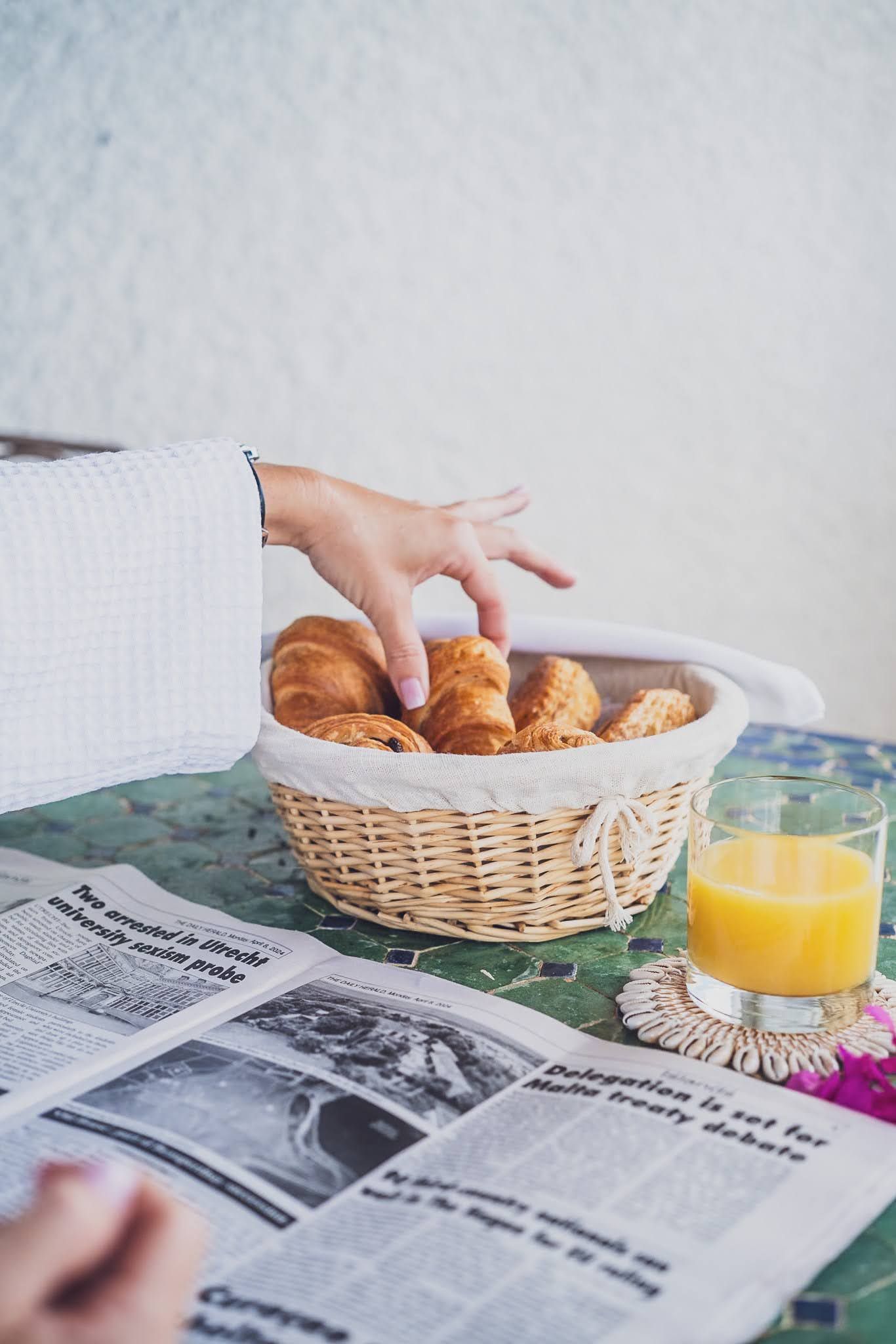 Hand reaching for croissant in basket, next to newspaper and orange juice.