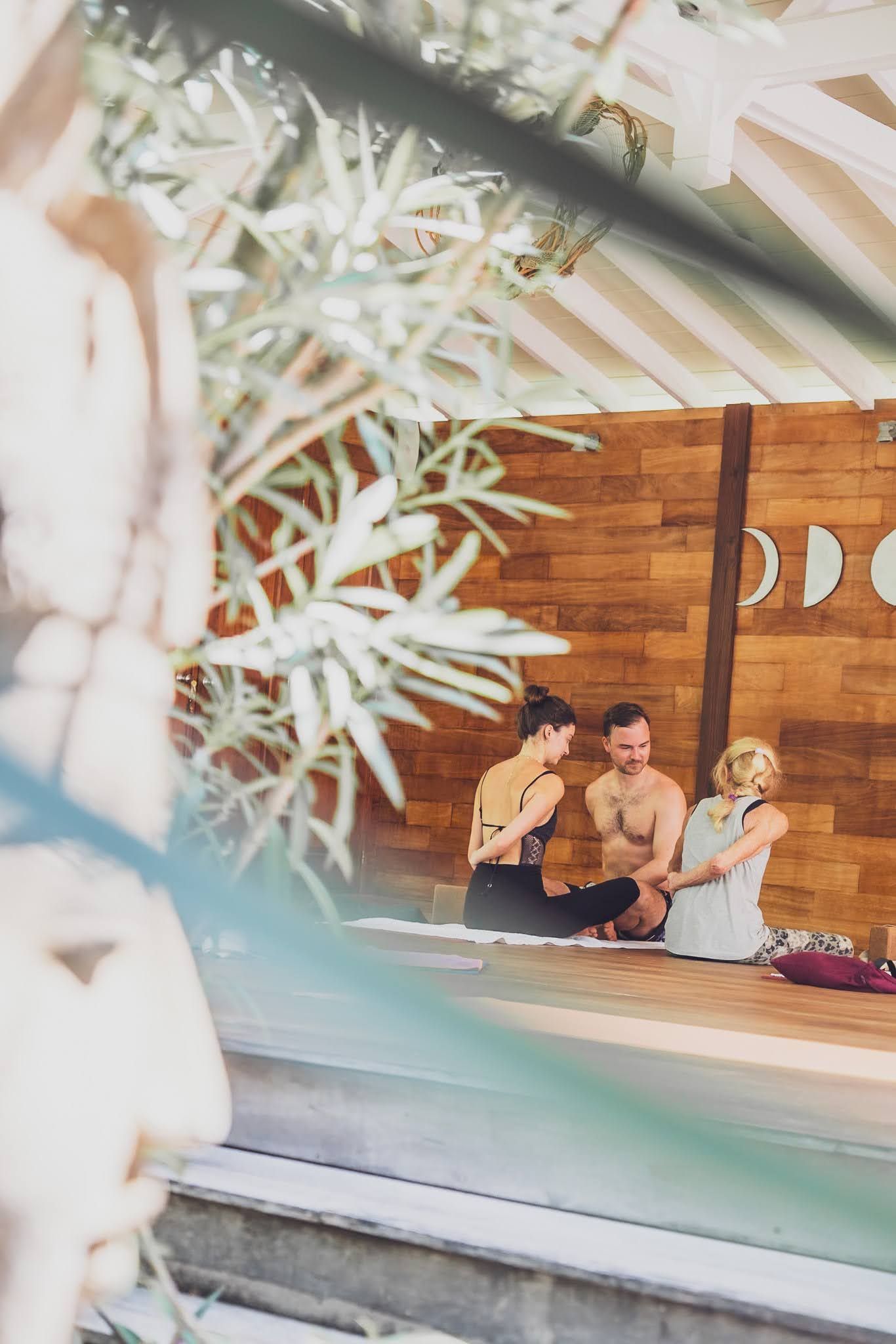Three people sitting on a wooden platform, engaged in a discussion. The setting is a bright, natural space.