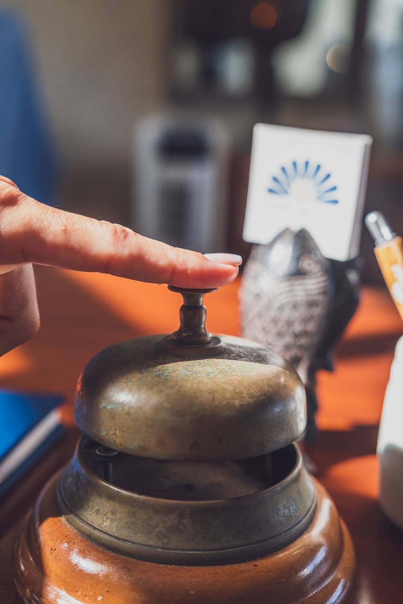 Finger pressing a brass service bell on a wooden desk, requesting assistance.