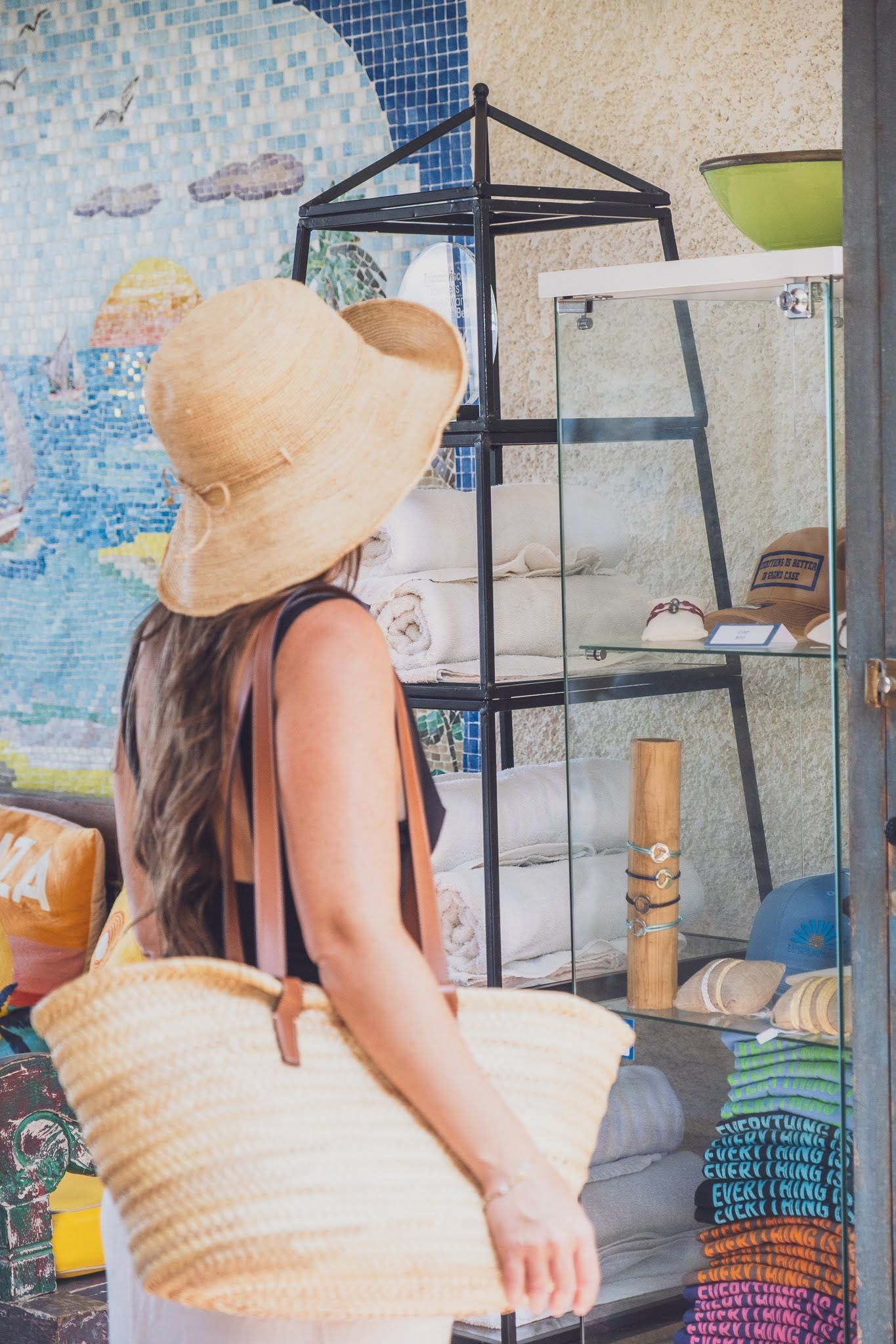Woman in straw hat looks at display case, holding a woven tote bag.