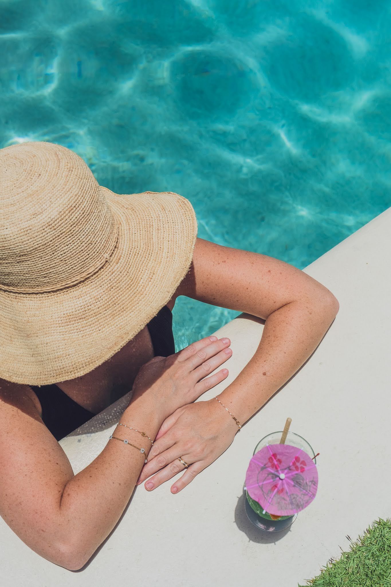 Woman in straw hat relaxing by a pool, arms resting on the edge, cocktail nearby.