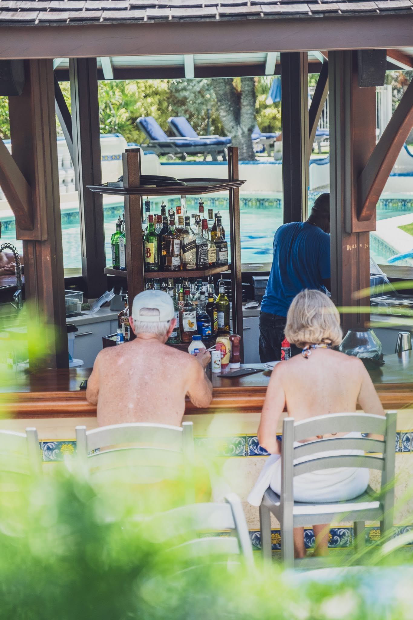 People at a poolside bar; bartender mixing drinks. Wooden structure, sunlight, tropical setting.