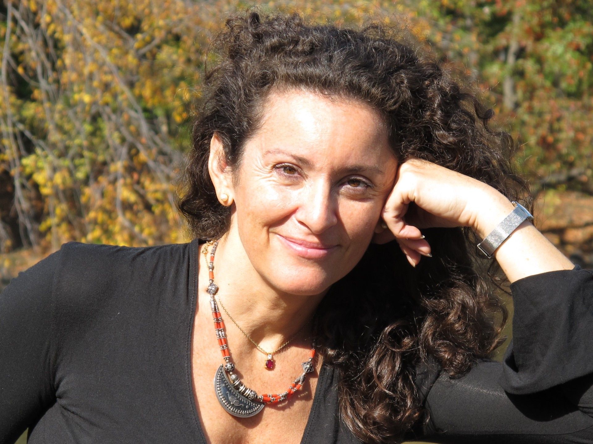 Woman with curly brown hair smiles, rests chin on hand, wearing black top and necklace.