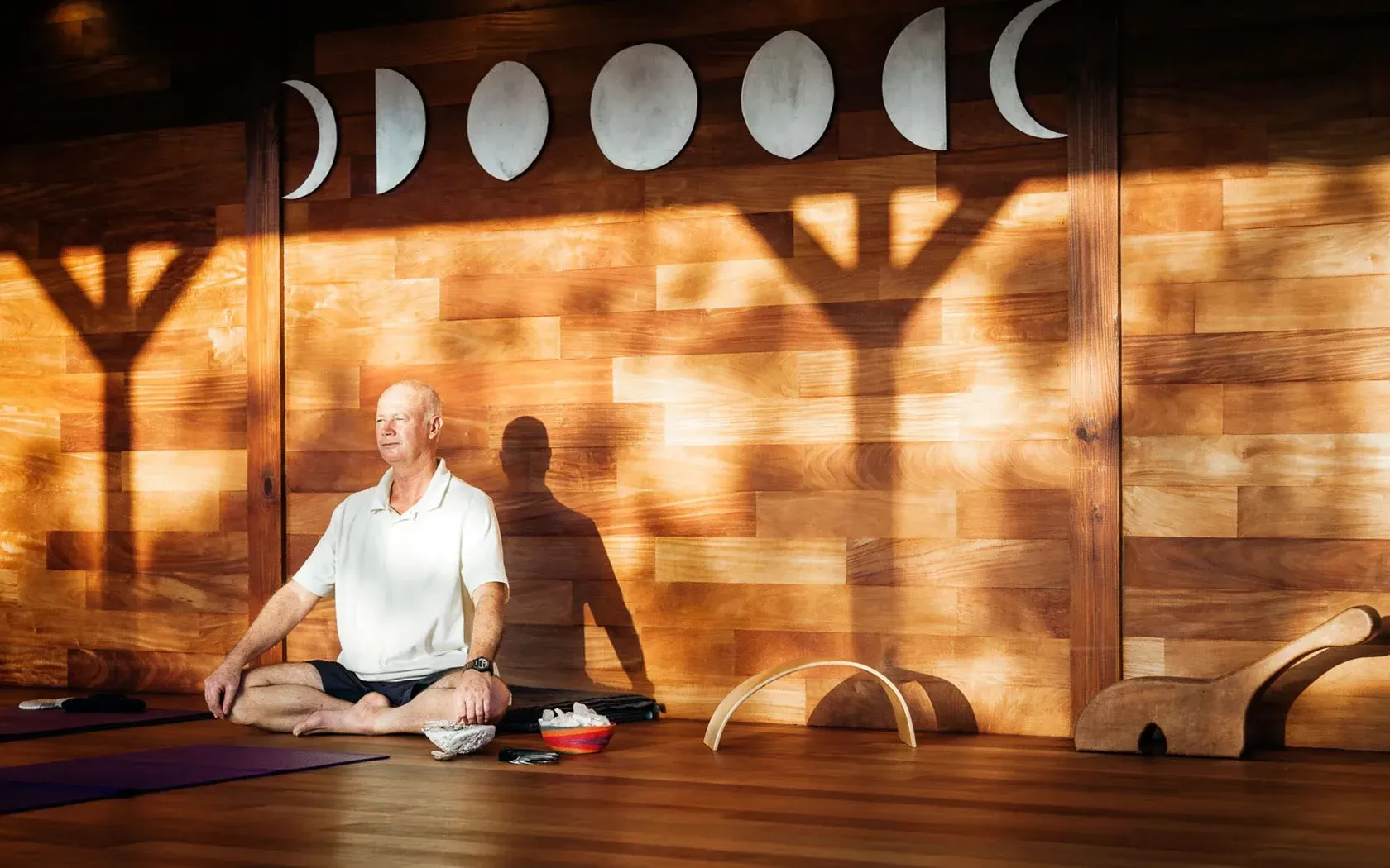 Man meditating in a wooden-walled room with moon phase art, sunlit shadows.