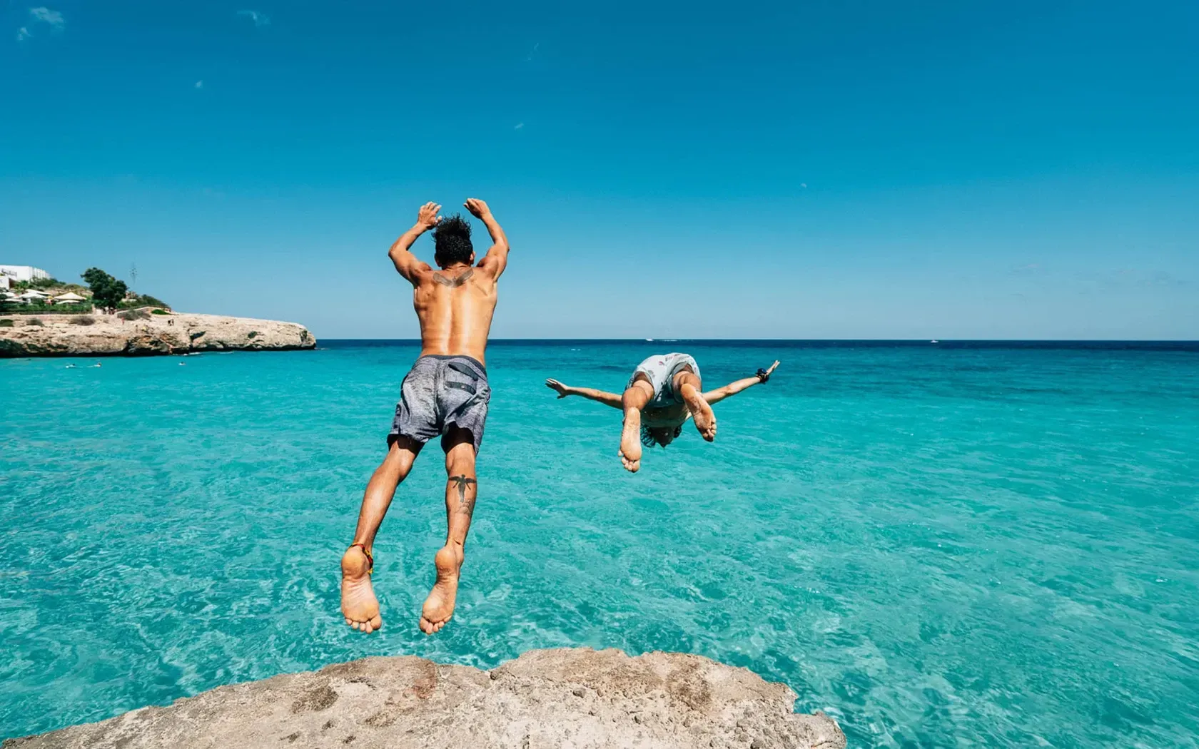 Two people jump into turquoise water from a rocky cliff under a blue sky.