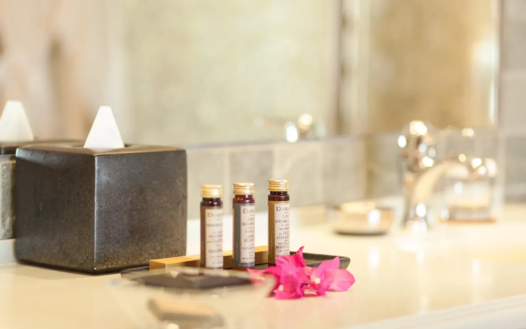 Bathroom counter with tissue box, small bottles, pink flower, and a blurred background.