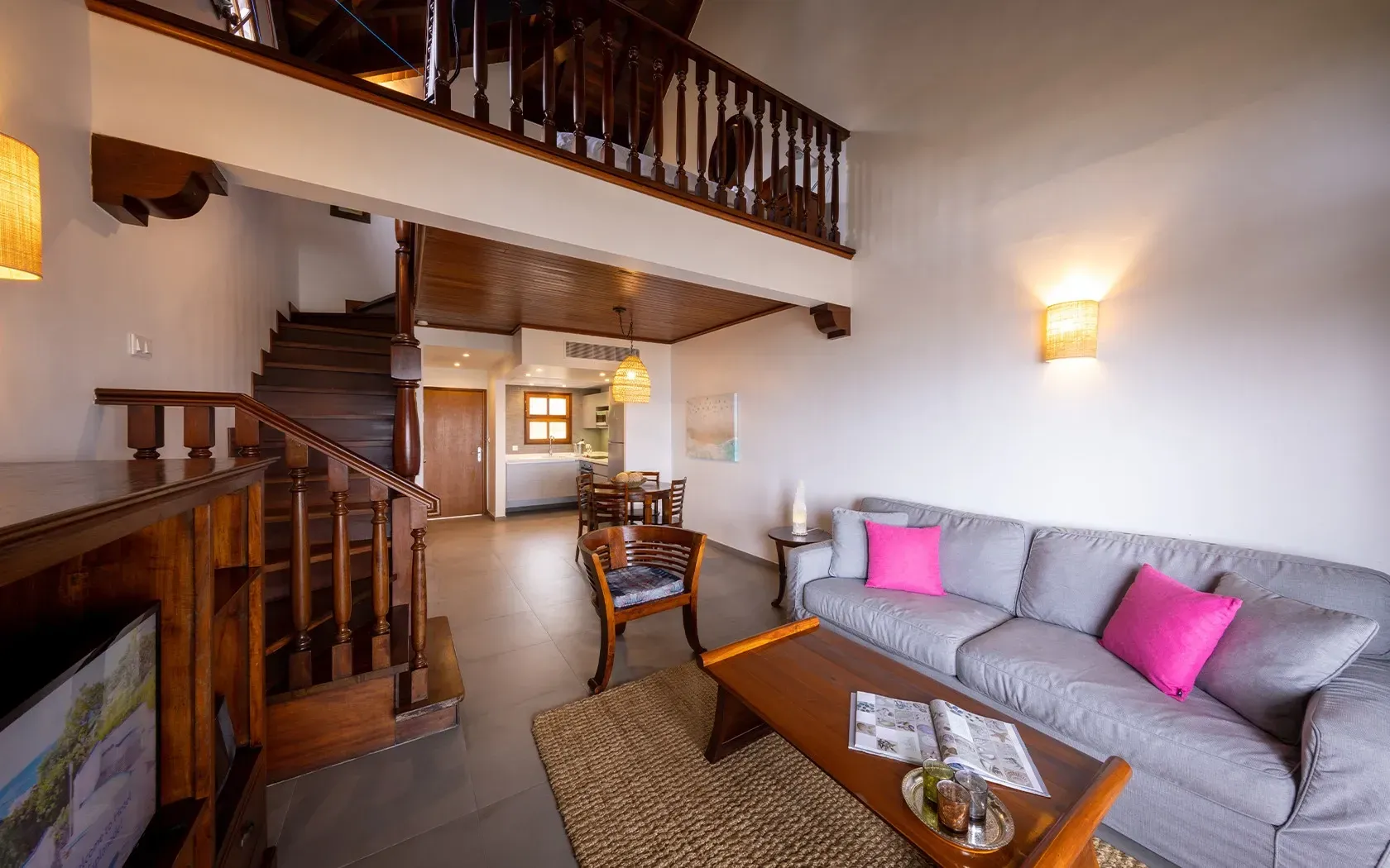 Two-story living room with wooden stairs and mezzanine. Gray sofa with pink pillows, brown coffee table, and TV.