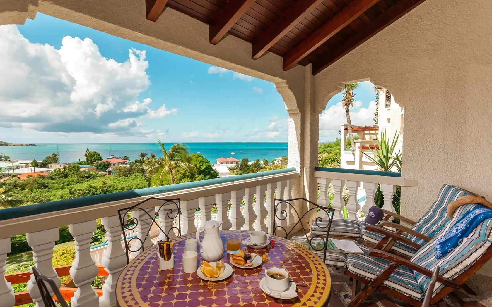 Balcony with table set for breakfast, overlooking ocean and lush greenery. Blue sky with fluffy clouds.