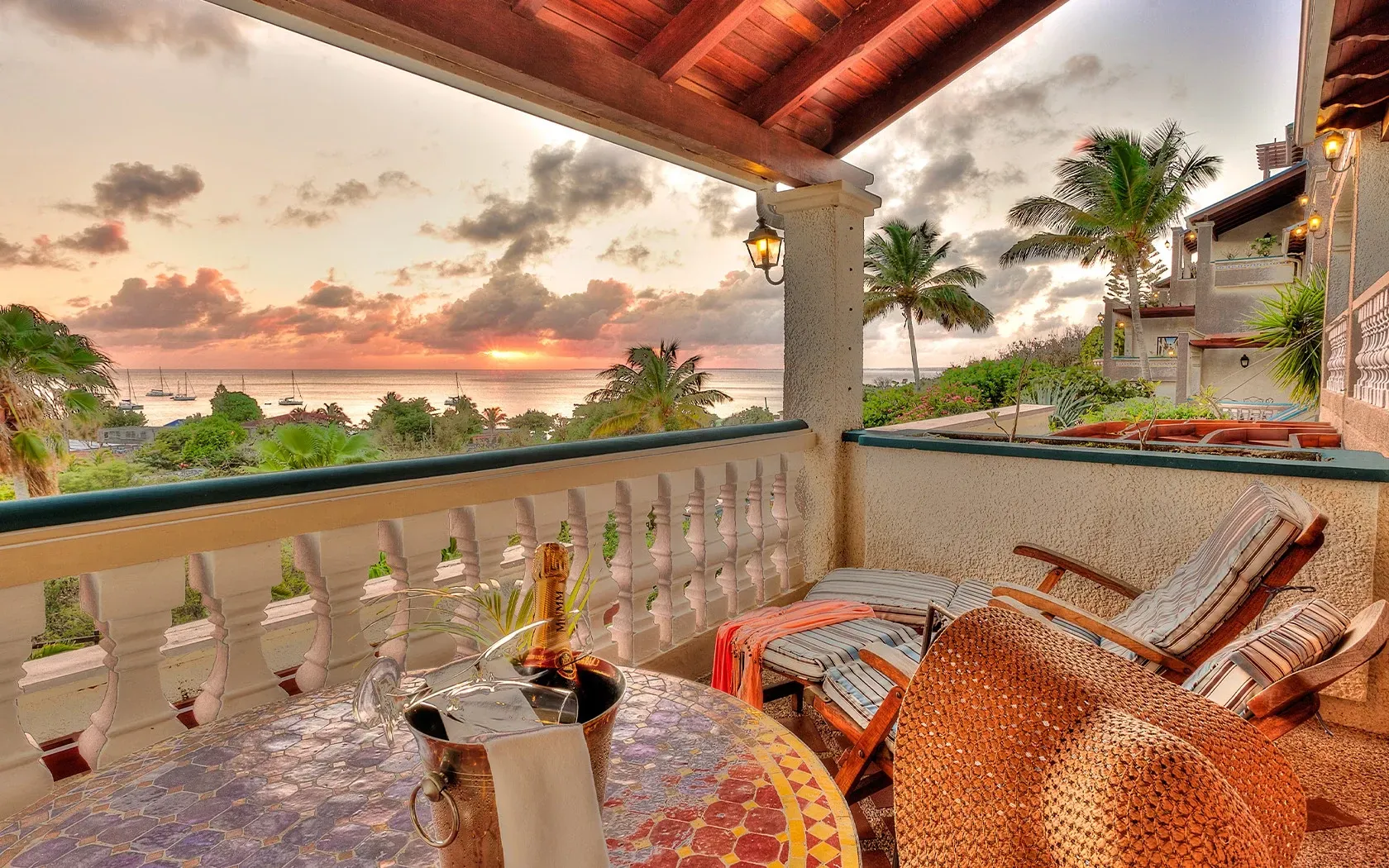 Balcony overlooking ocean at sunset; table, chairs, tropical foliage, and a stucco building.