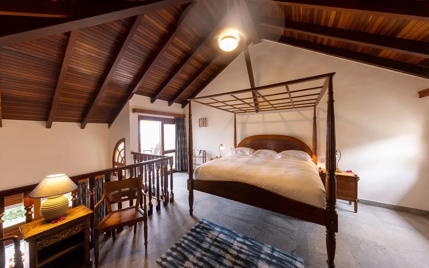 Bedroom with a four-poster bed under a wooden sloped ceiling; small balcony visible, warm lighting.
