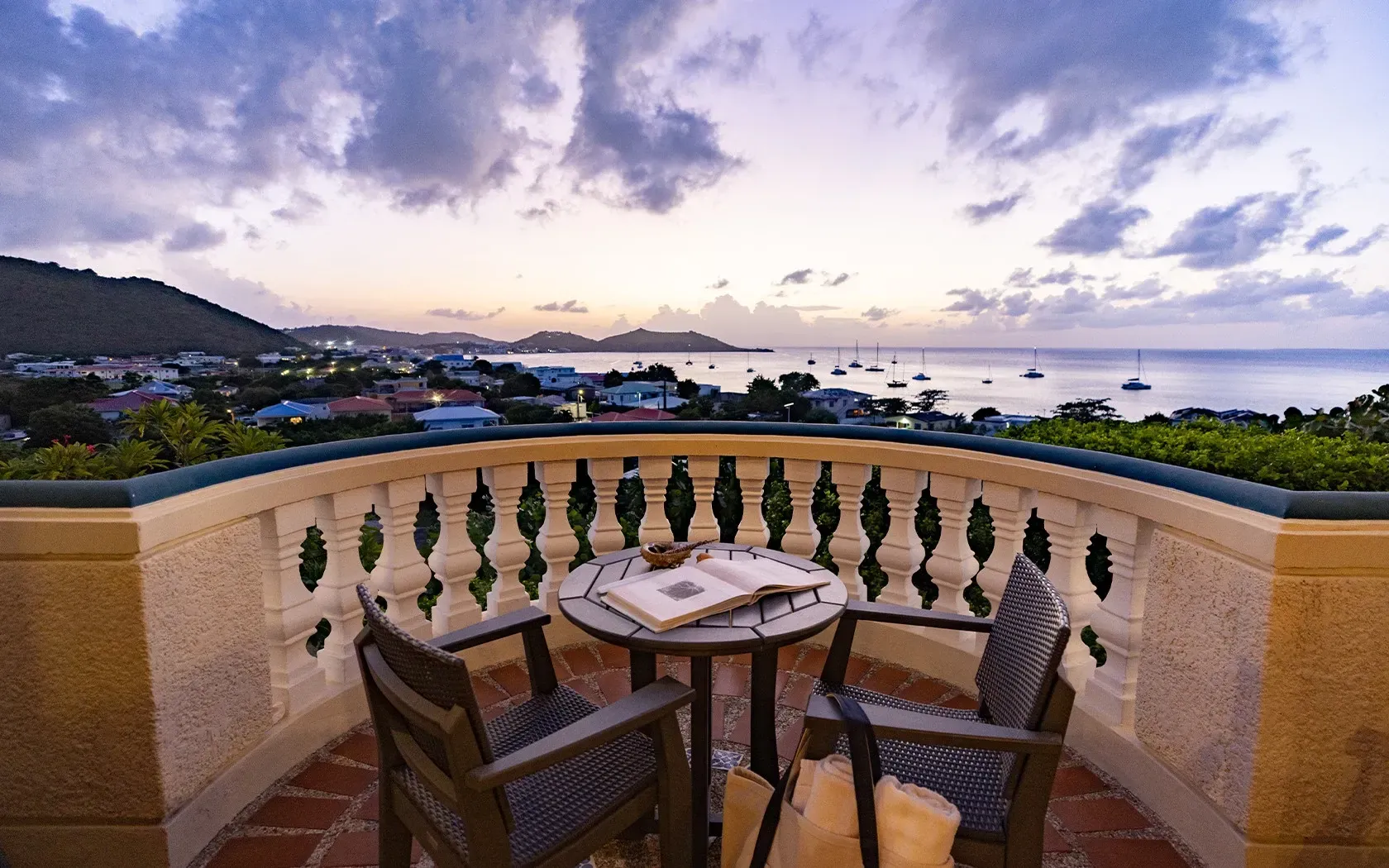 Balcony with table and chairs overlooking ocean bay at dusk.
