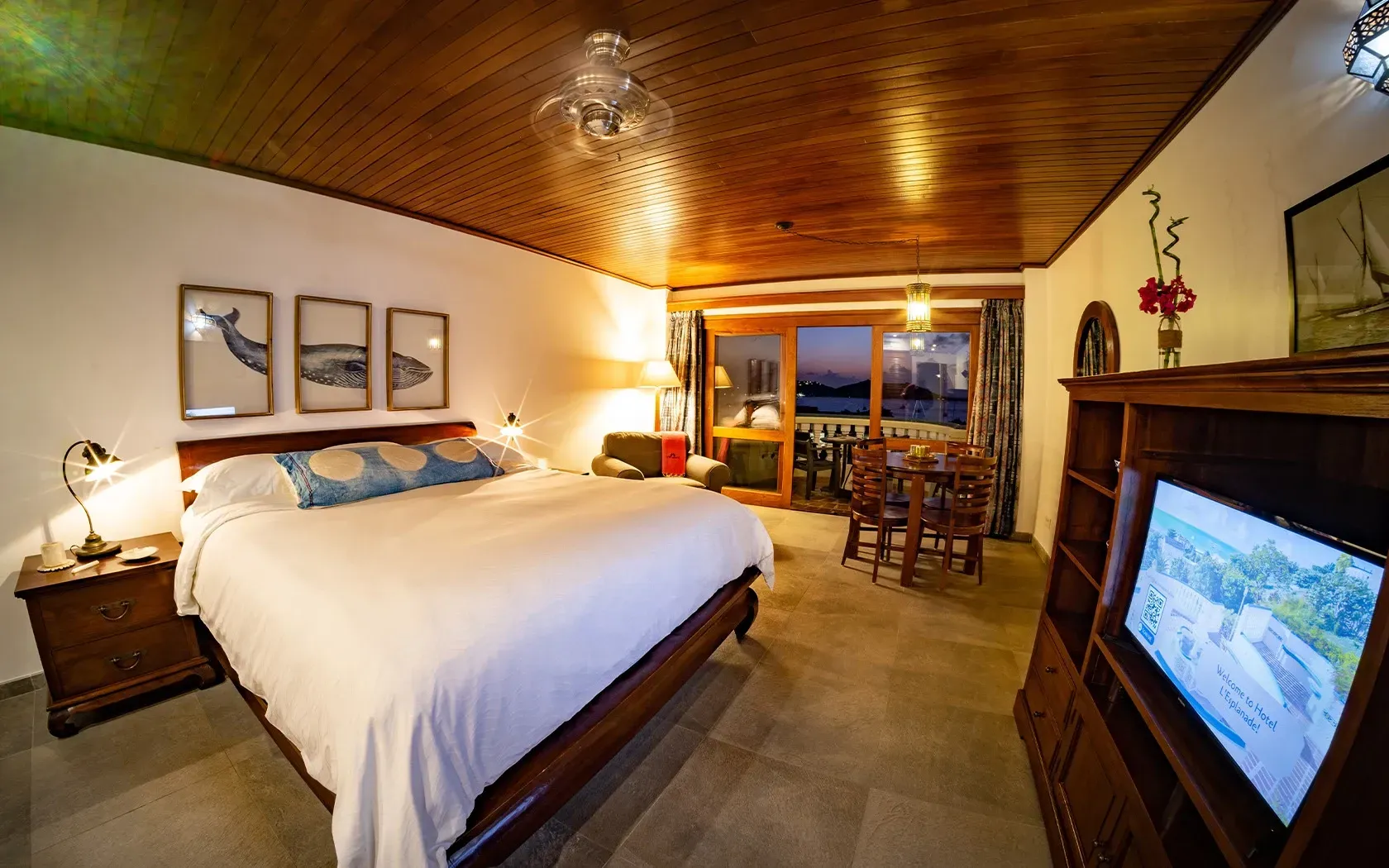 Cozy hotel room: bed, ocean view balcony, wood ceiling, artwork, TV, brown furniture.