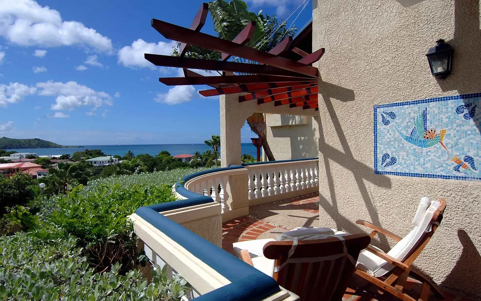 Balcony overlooking ocean with pergola, blue railing, mosaic art, and wooden chairs.