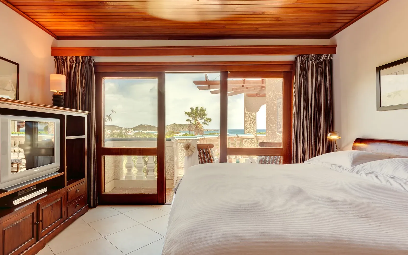 Bedroom with large bed, wooden ceiling, balcony view of ocean and palm tree.