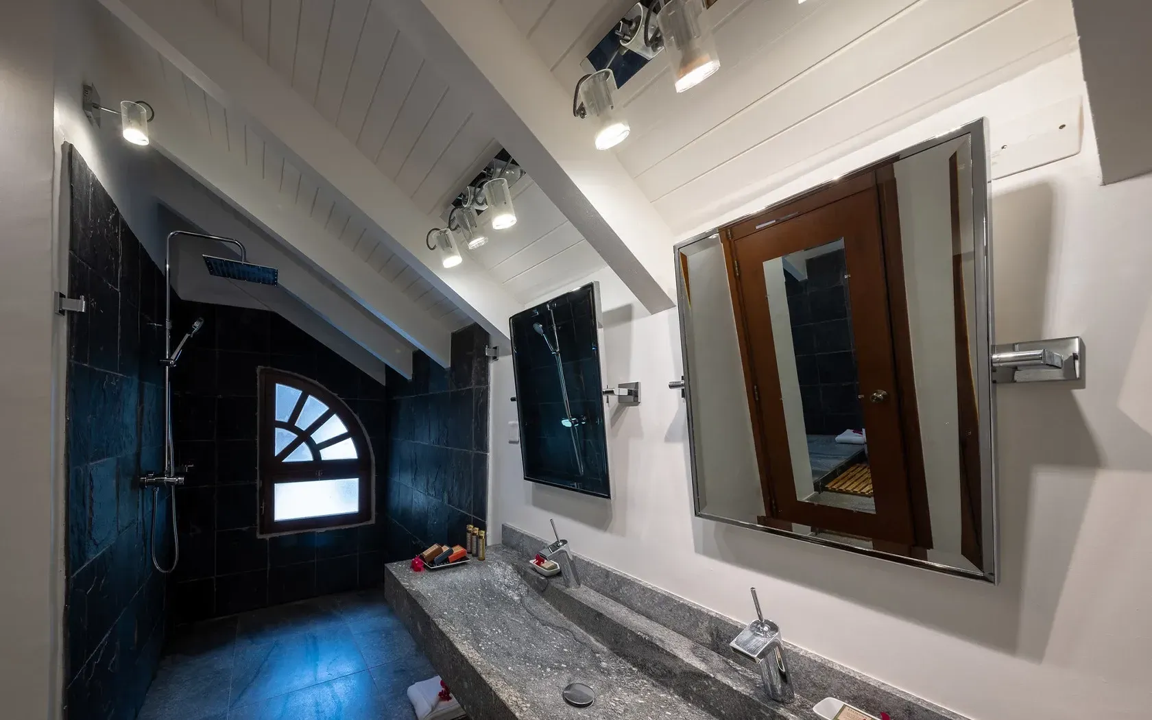 Bathroom with angled ceiling, dual sinks, black tiled shower, arched window, and mirrors.