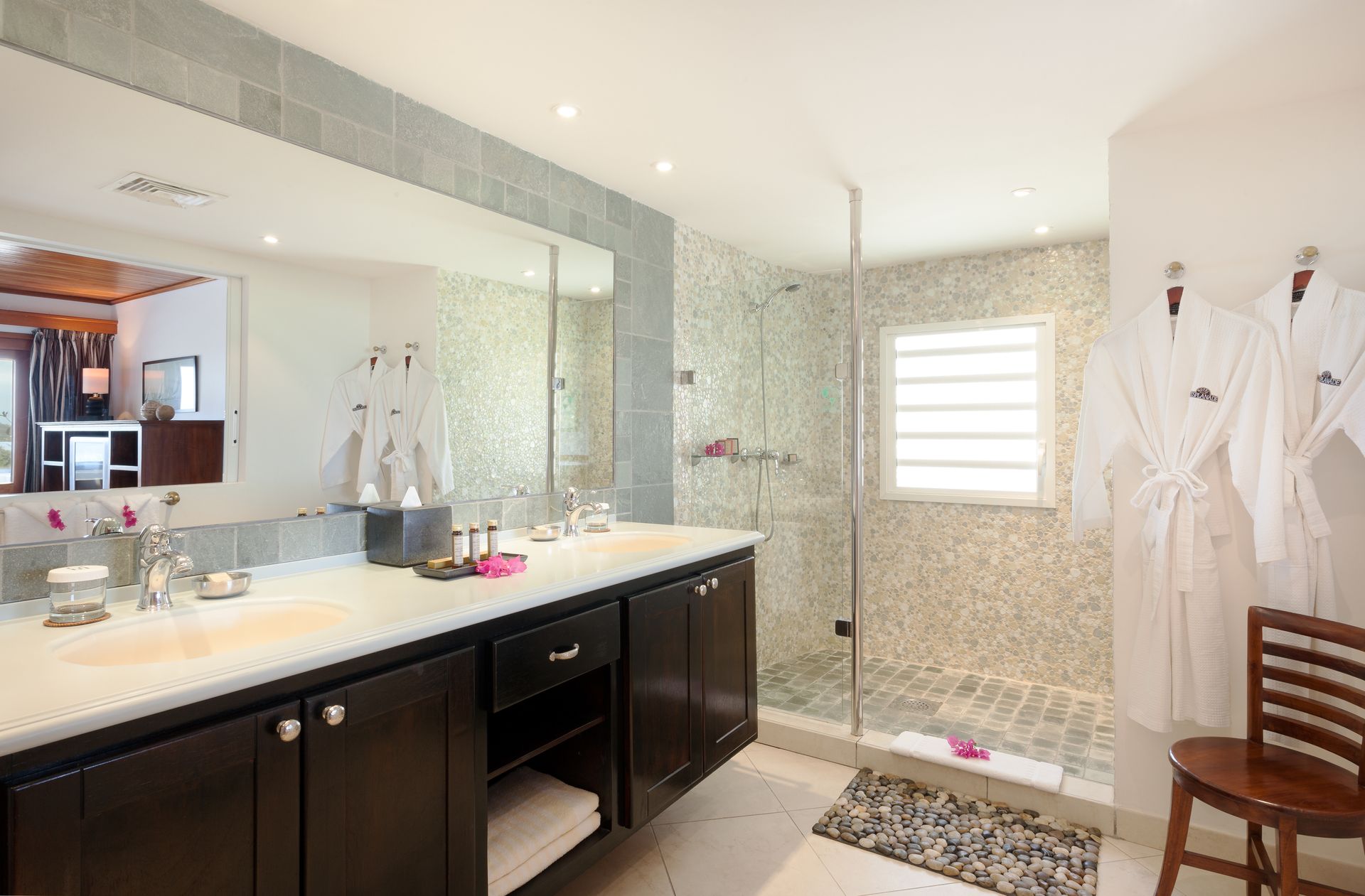 Modern bathroom with dark cabinets, a large mirror, and a glass shower. White robes hang on the wall.