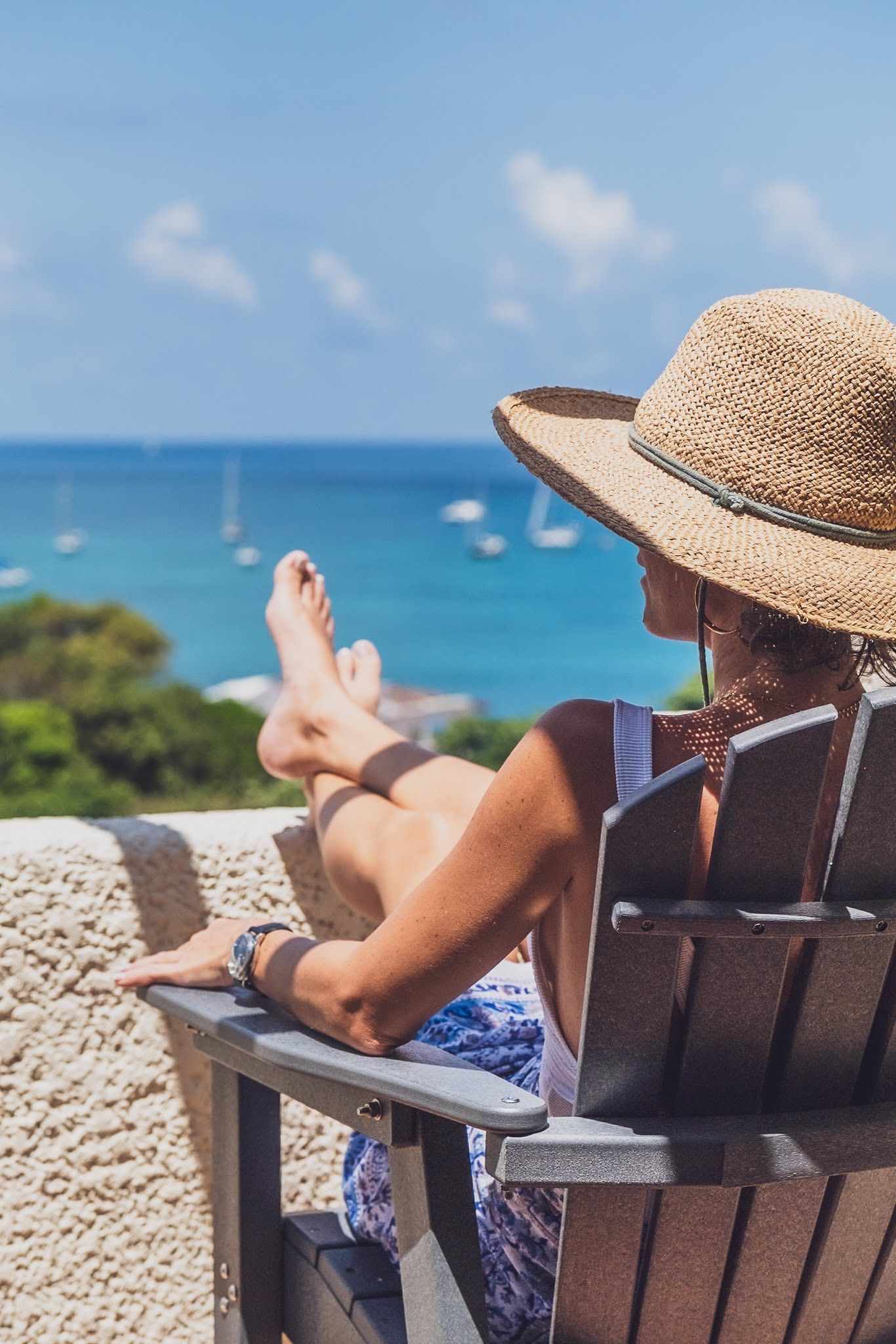 Woman in sun hat relaxes in chair, looking at ocean view with sailboats.