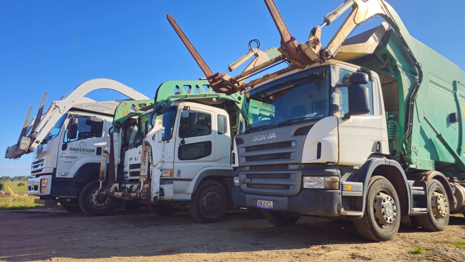 Three garbage trucks parked side-by-side outdoors under a blue sky, each with a different color cab and lift mechanism.